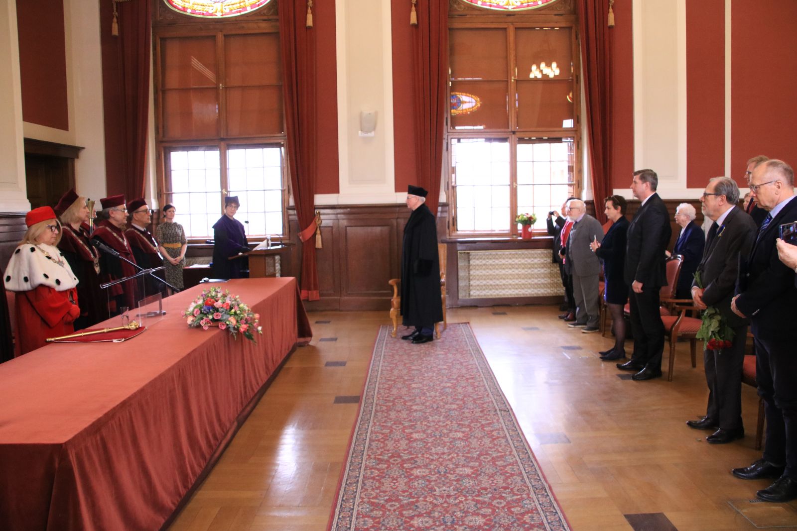 Prof. Danielewicz stoi pomiędzy stołem prezydialnym, zwrócony do grona rektorskiego, a publicznością. Wszyscy stoją.