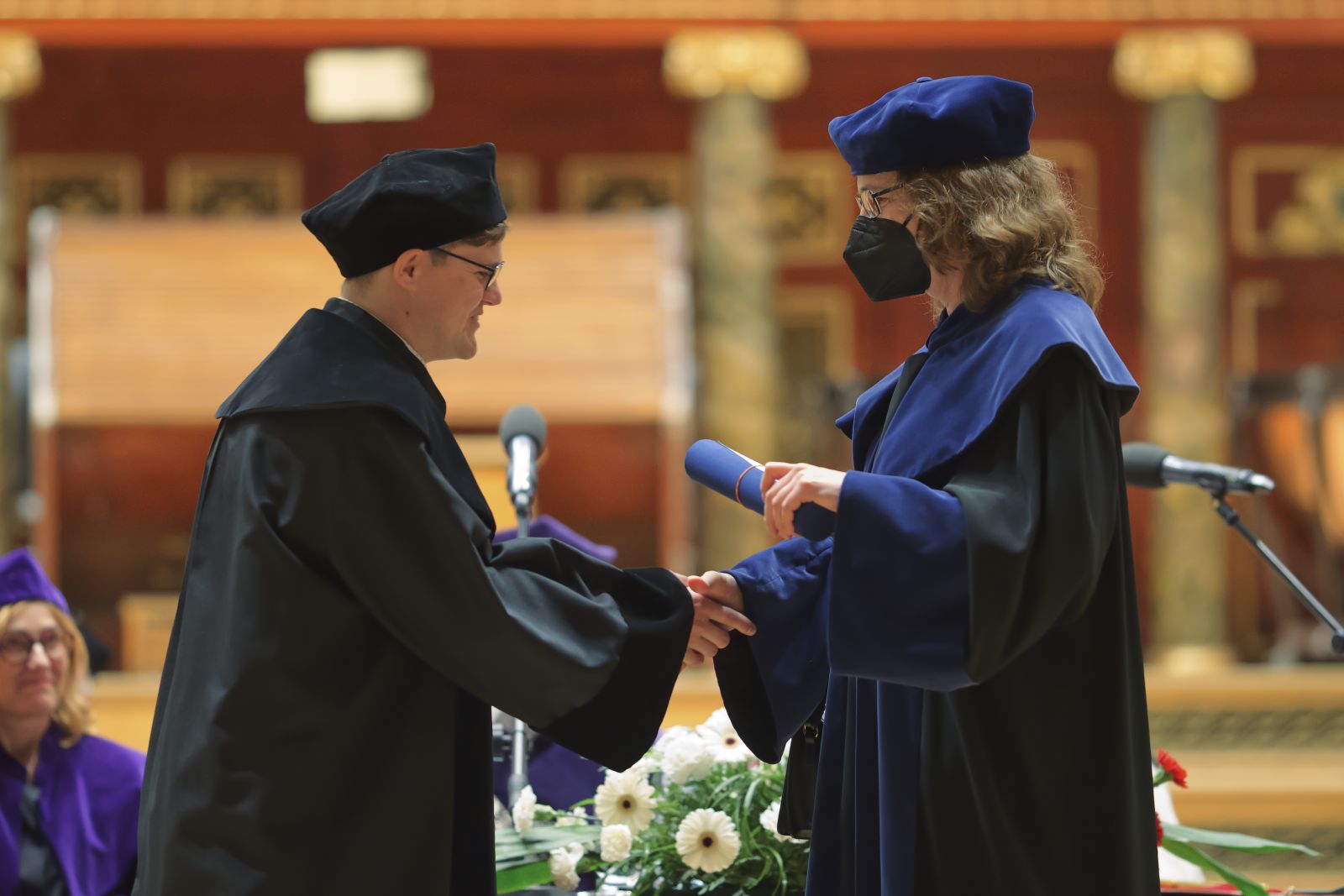 Dwie osoby w togach, kobieta w maseczce na twarzy wręcza mężczyźnie dyplom.