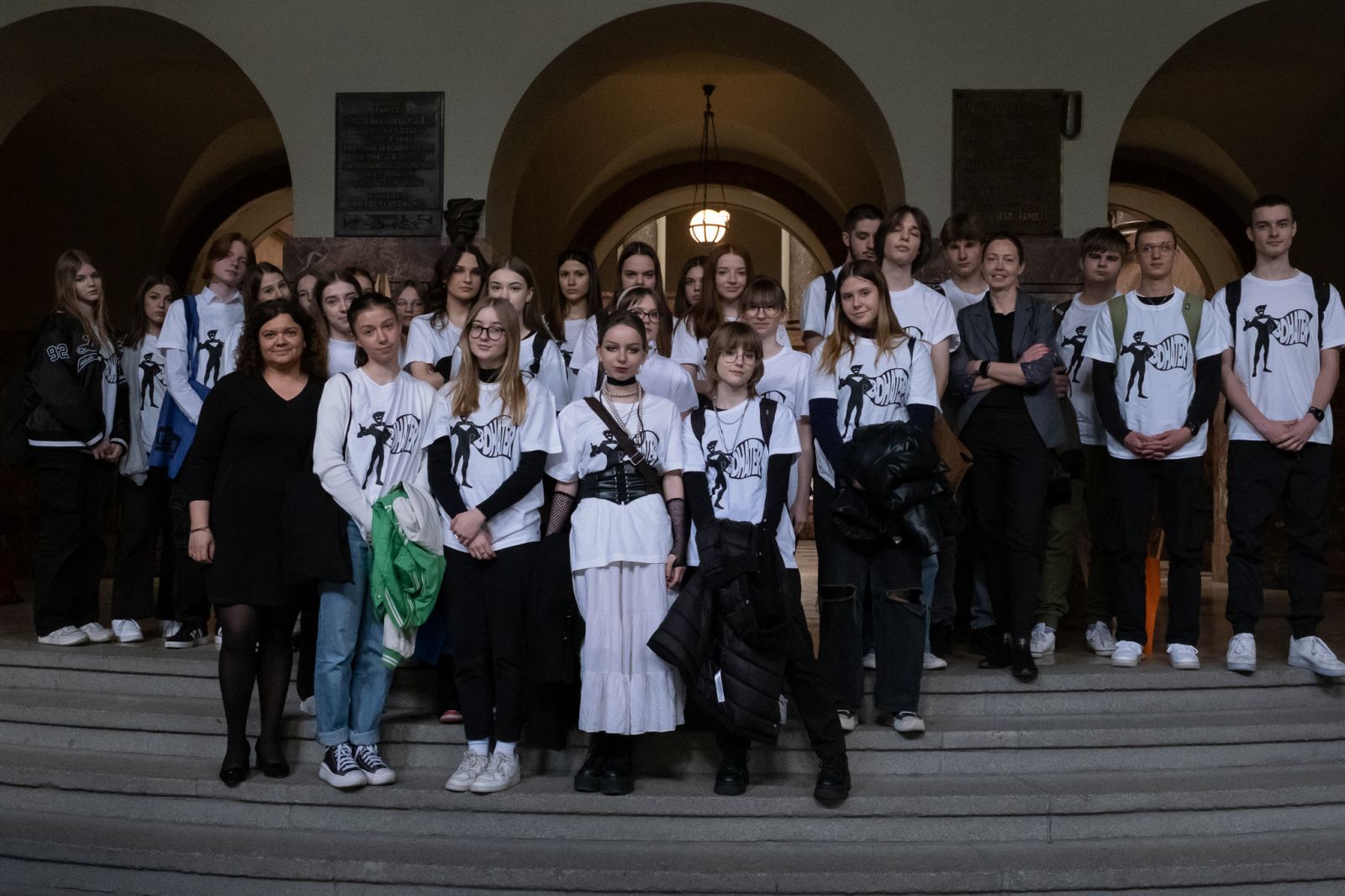 Zdjęcie grupowe kilkudziesięciu młodych ludzi ubranych w identyczne białe koszulki, stojących rzędami na schodach w holu głównym Collegium Maius.
