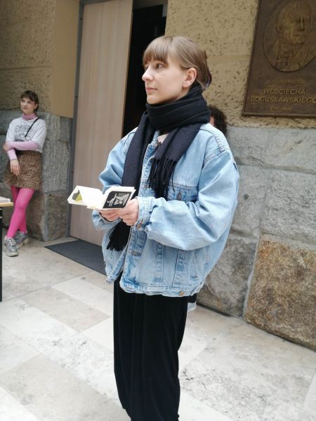 W holu biblioteki WFPiK stoi młoda kobieta, w dłoniach trzyma otwartą małą książkę.
