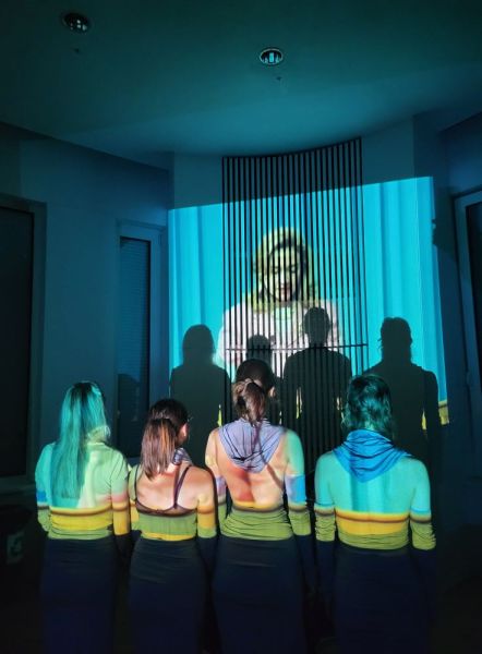 Zdjęcie z przedstawienia teatru Sfinga. 4 kobiety stoją tyłem do obiektywu, na ich plecy skierowane jest światło rzutnika, które na ścianę na przeciwko rzuca obraz młodej blondynki oraz cienie sylwetek 4 kobiet.