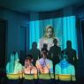 Zdjęcie z przedstawienia teatru Sfinga. 4 kobiety stoją tyłem do obiektywu, na ich plecy skierowane jest światło rzutnika, które na ścianę na przeciwko rzuca obraz młodej blondynki oraz cienie sylwetek 4 kobiet.