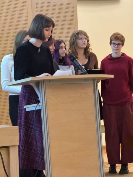 Przy mównicy w Salonie Mickiewicza stoi uczennica i odczytuje swoje wystąpienie, za nią stoją inni uczniowie i uczennice.