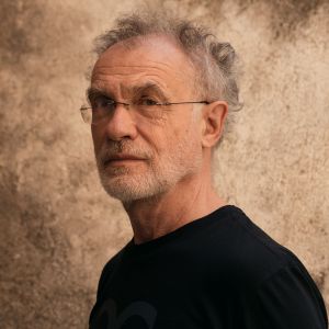 Zdjęcie starszego mężczyzny w czarnej koszulce, okularach, z brodą i przerzedzonymi, kręcącymi się siwymi włosami, patrzącego w obiektyw z nieodgadnioną miną.