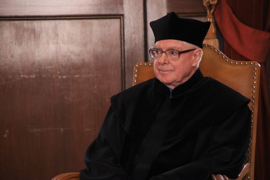 Prof. Jerzy Danielewicz siedzi na ozdobnym krześle, ubrany w czarną togę i biret.