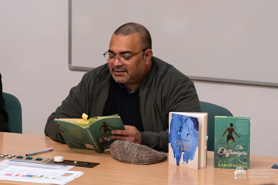 Sabin Iqbal siedzi przy stole, czyta z otwartej książki, przed nim stoją jeszcze dwie inne książki - jedna o polskim tytule 
