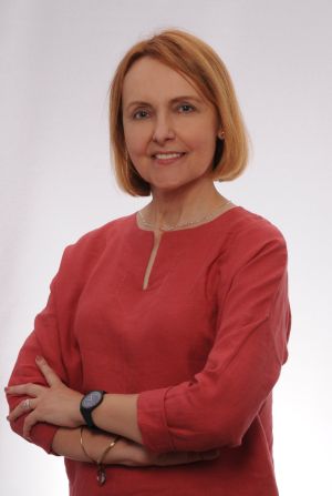 Zdjęcie kobiety w średnim wieku, o blond włosach, w czerwonej bluzce i srebrnym łańcuszku na szyi, stoi ze skrzyżowanymi na piersiach rękoma, uśmiecha się lekko do obiektywu.