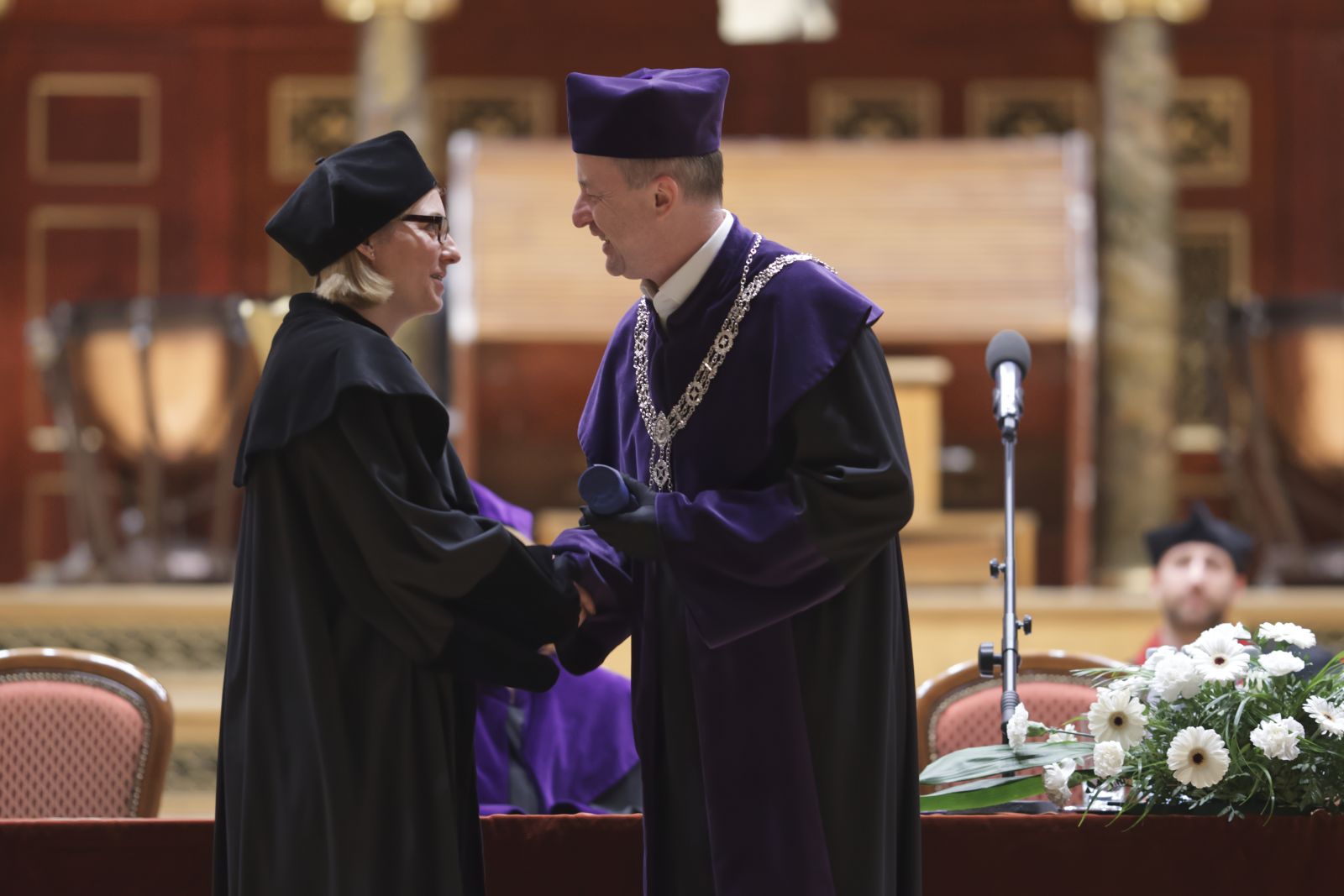 Dziekan Tomasz Mizerkiewicz ściska dłoń kobiety ubranej w czarną togę, wręczając jej dyplom.