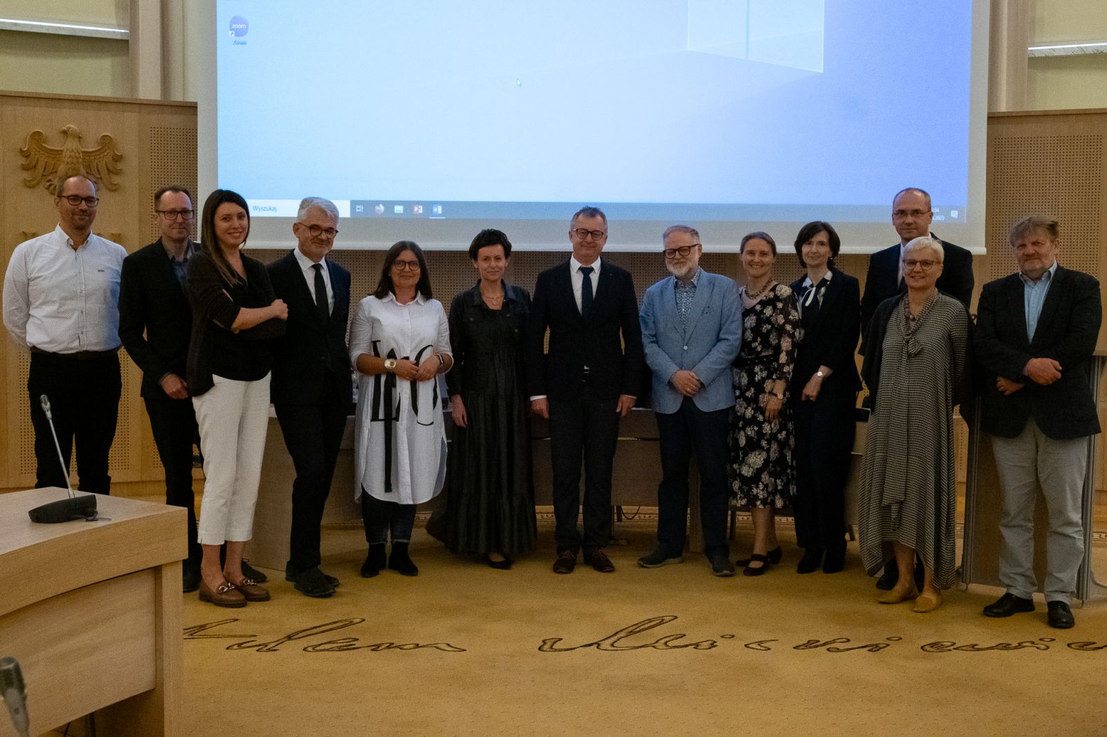 Zdjęcie grupy 13 osób w średnim i starszym wieku, elegancko ubranych, stojących pośrodku Salonu Mickiewicza pod Kopułą Collegium Maius.