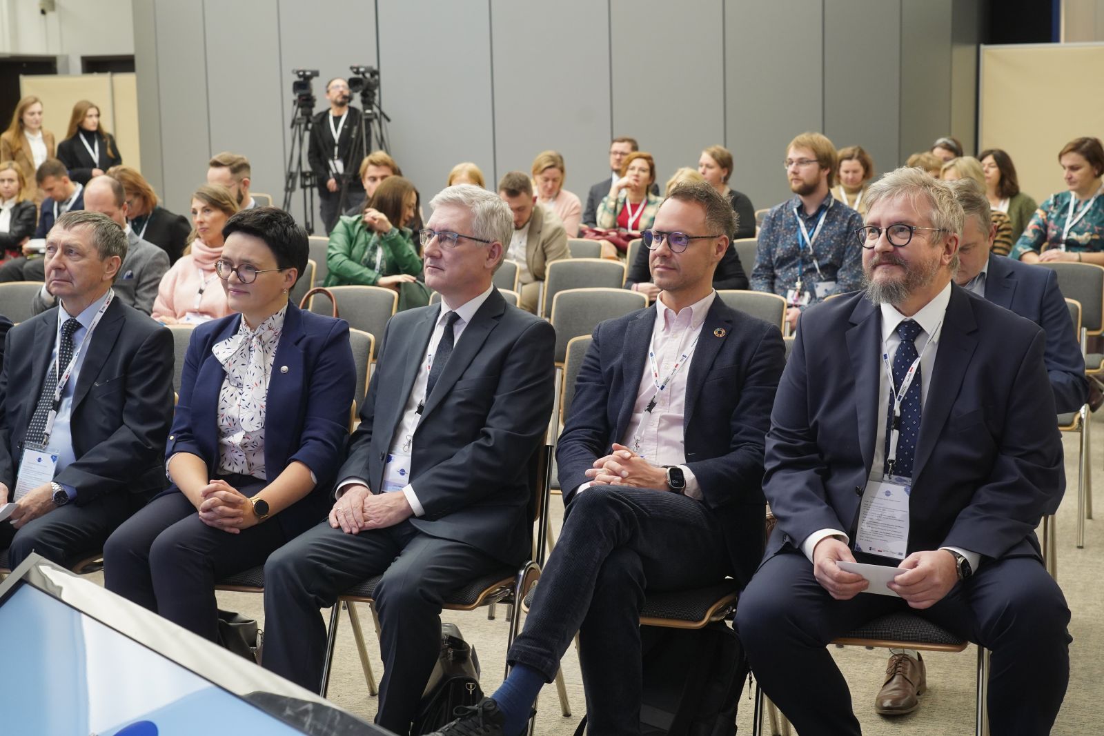 W dużej sali konferencyjnej o nowoczesnym wyposażeniu widać rzędy krzeseł, na których siedzi kilkadziesiąt osób. Z tyłu widać dwie kamery i kamerzystę. Na pierwszym planie widać pięć osób (4 mężczyzn i 1 kobietę) siedzących na krzesłach, patrzących nieco w bok od obiektywu, zasłuchanych.