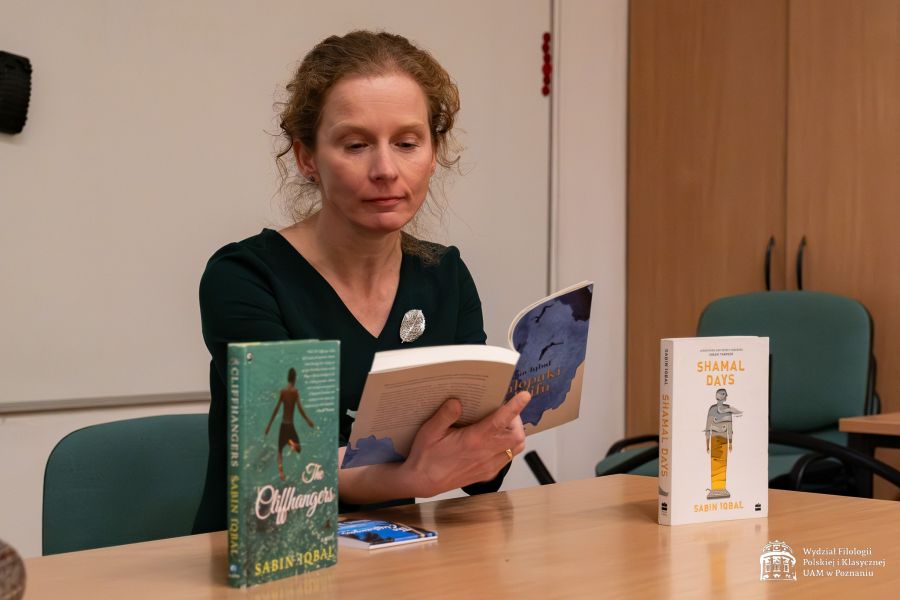 Prof. Ewa Rajewska siedzi przy stole czyta z otwartej książki, przed nią stoją jeszcze dwie inne książki Sabina Iqbala z tytułami w języku angielskim.