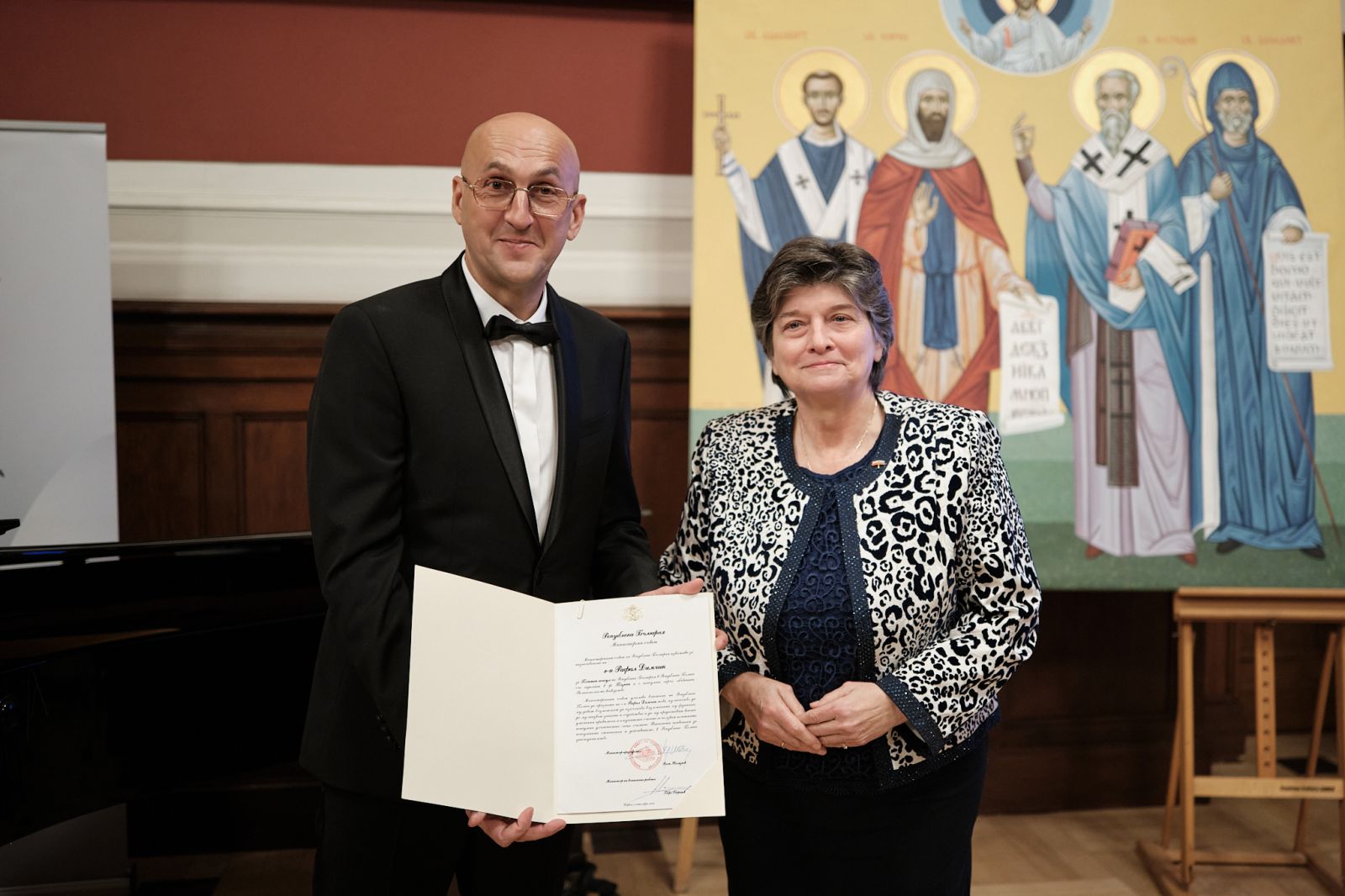 Nowy konsul i Ambasador Bułgarii prezentują do obiektywu akt mianowania, za nimi na sztalugach stoi prawosławna ikona.