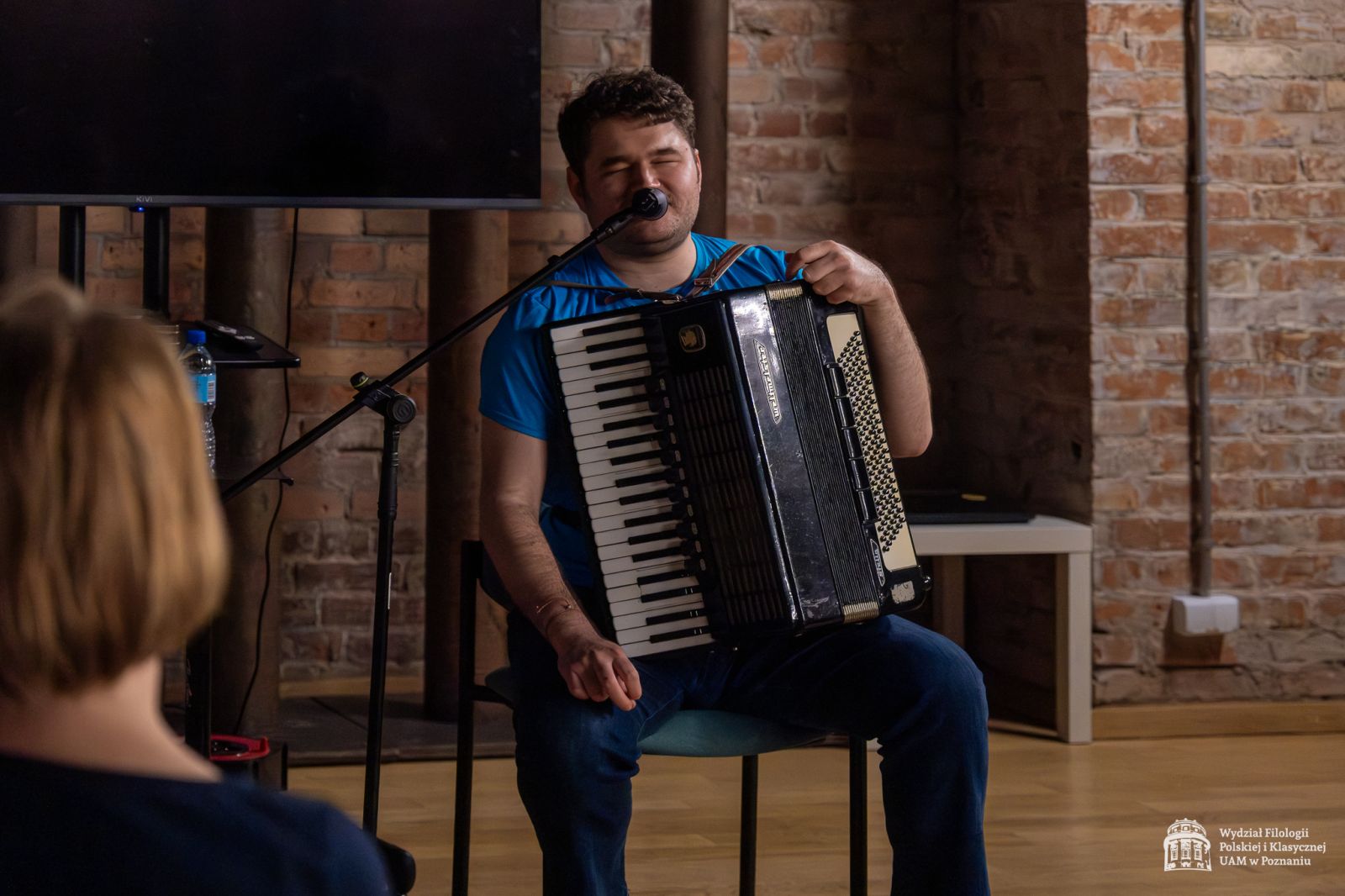 Mężczyzna z akordeonem siedzi w Sali Teatralnej i mówi do mikrofonu. Ma zamknięte oczy, jest niewidomy.