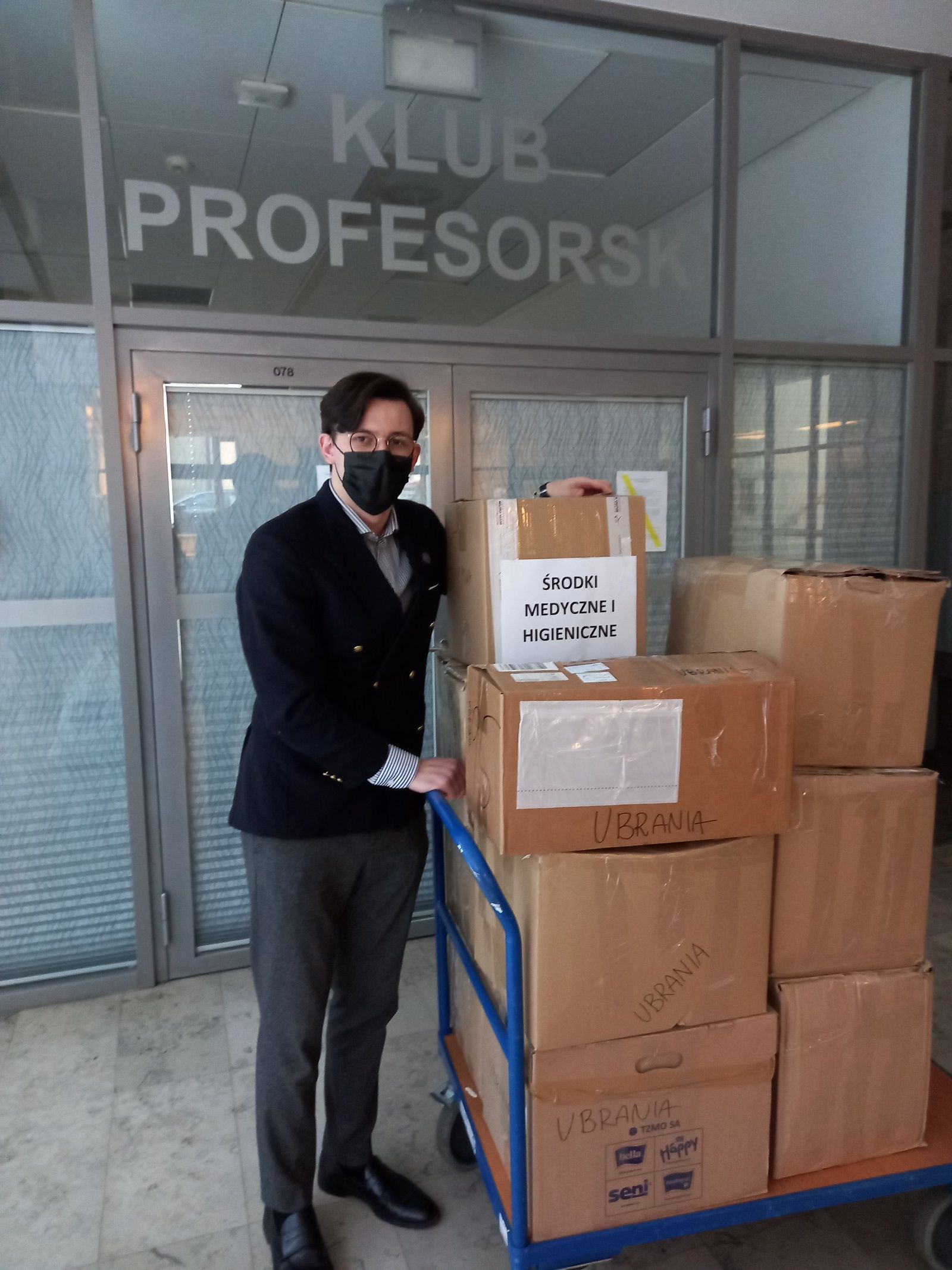 Przemysław Raczyk z Rady Samorządu Studentów WFPiK stoi obok wózka towarowego załadowanego kartonami z darami dla Ukrainy. Kartony sięgają mu prawie do głowy. Na kartonach napisy w rodzaju 