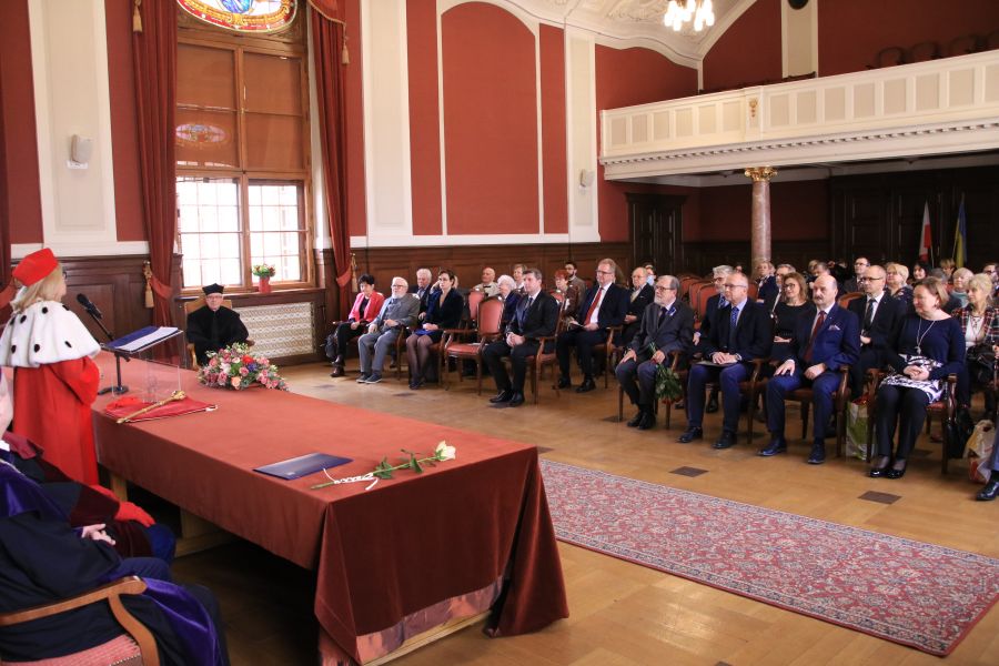Widok stołu prezydialnego, za którym siedzi Rektor UAM w czerwonej todze i płaszczu z gronostajów, oraz na widownię w Auli Lubrańskiego.