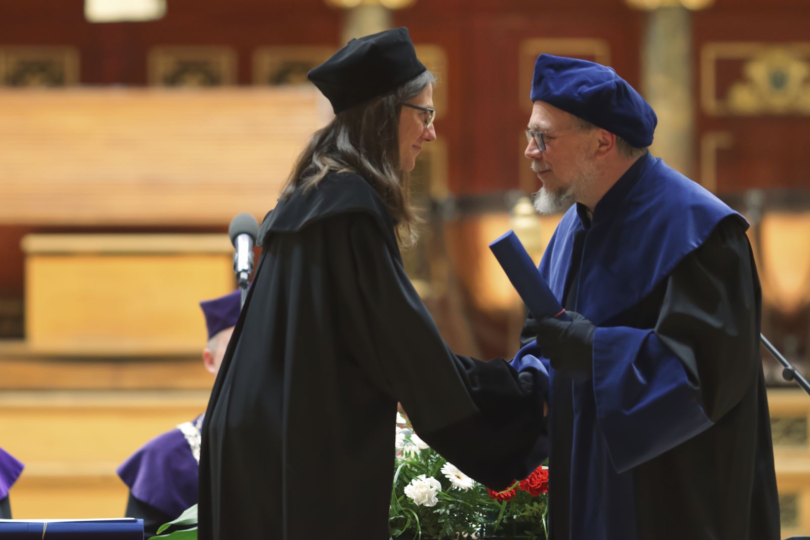 Dwie osoby w togach, starszy mężczyzna w okularach wręcza młodej kobiecie dyplom, ściskając jej dłoń.