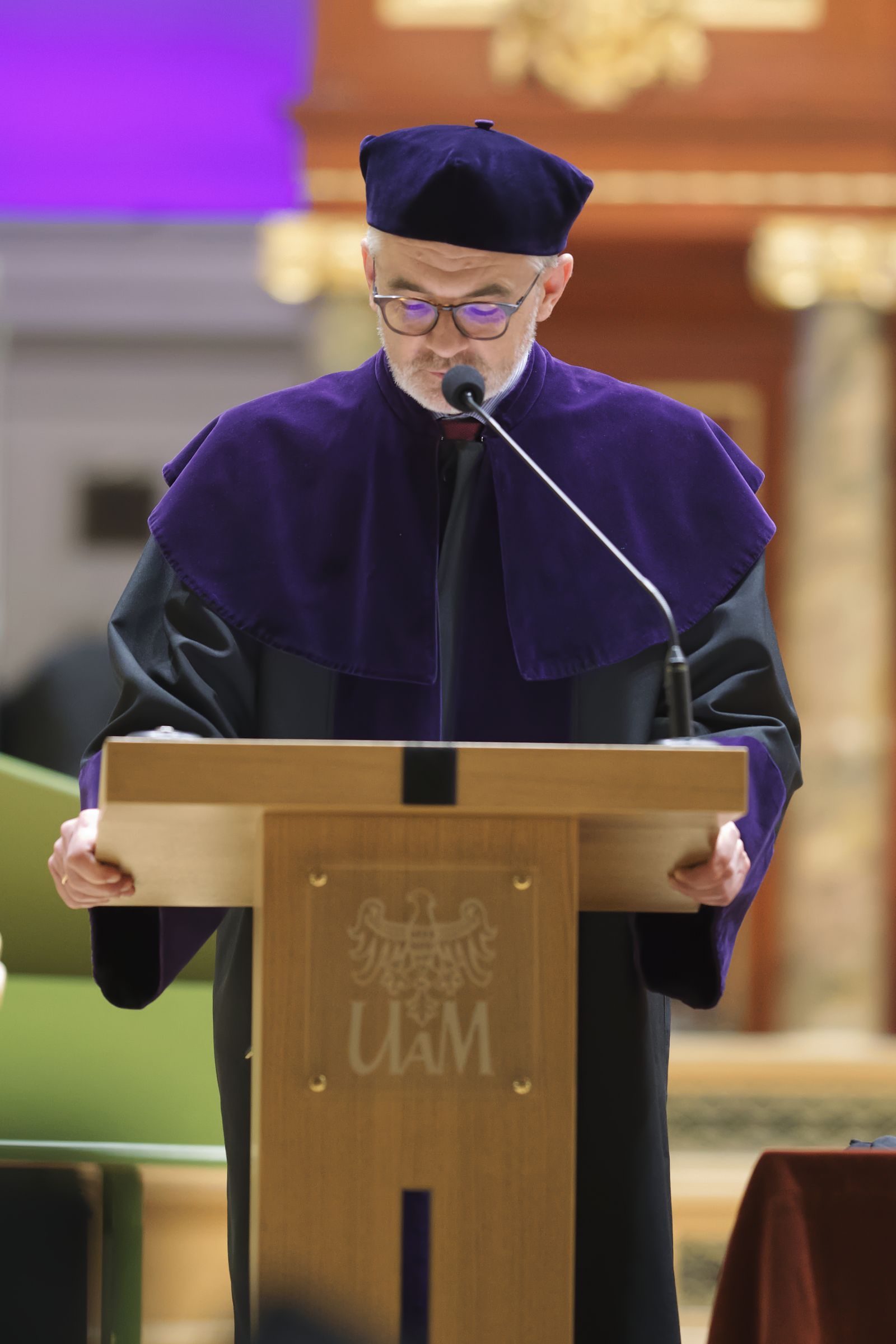 Prof. Jerzy Borowczyk stoi na mównicy w auli uniwersyteckiej i przemawia do mikrofonu, ubrany jest w fioletową togę.