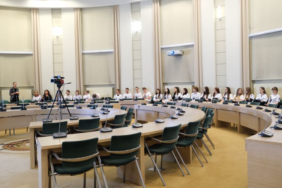 Grupa kilkudziesięciu młodych osób w identycznych białych koszulkach siedzi na krzesłach ustawionych w okręgu pod ścianą Salonu Mickiewicza pod Kopułą Collegium Maius. Większość z nich nie patrzy w obiektyw. Na środku Salonu ustawiona jest kamera.
