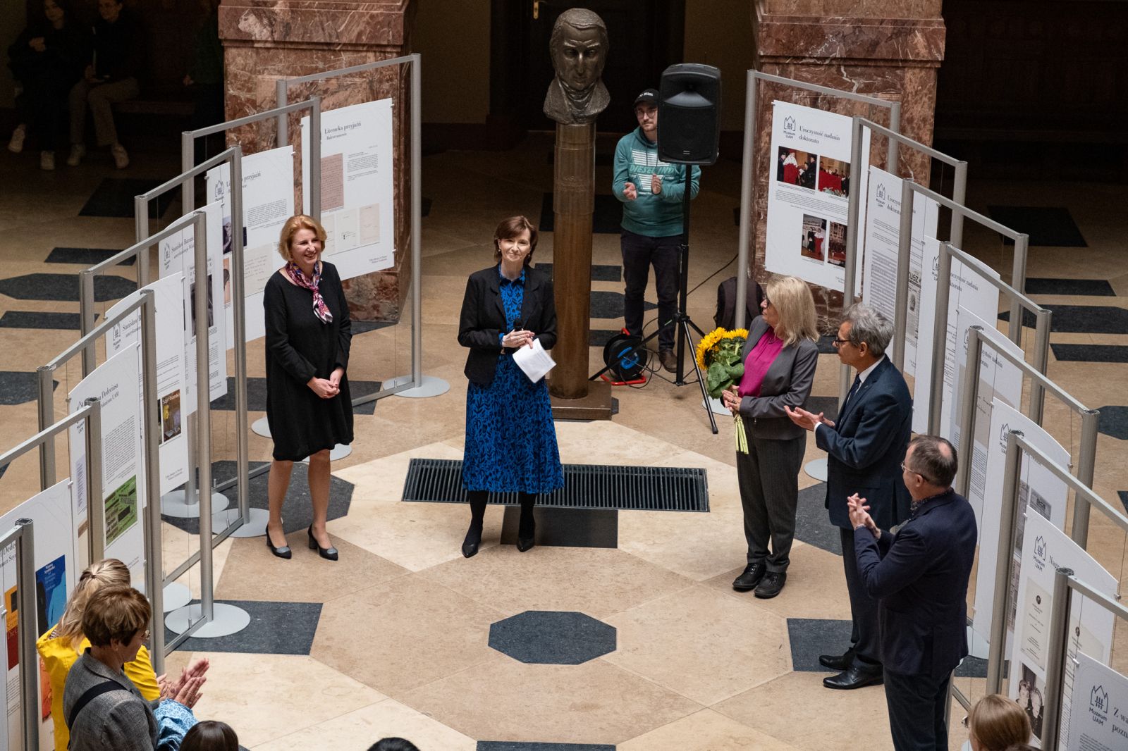 Zdjęcie z góry holu głównego Collegium Maius, gdzie stoi wystawa o Wisławie Szymborskiej. Przed planszami stoją uczestnicy wernisażu, klaszczą.