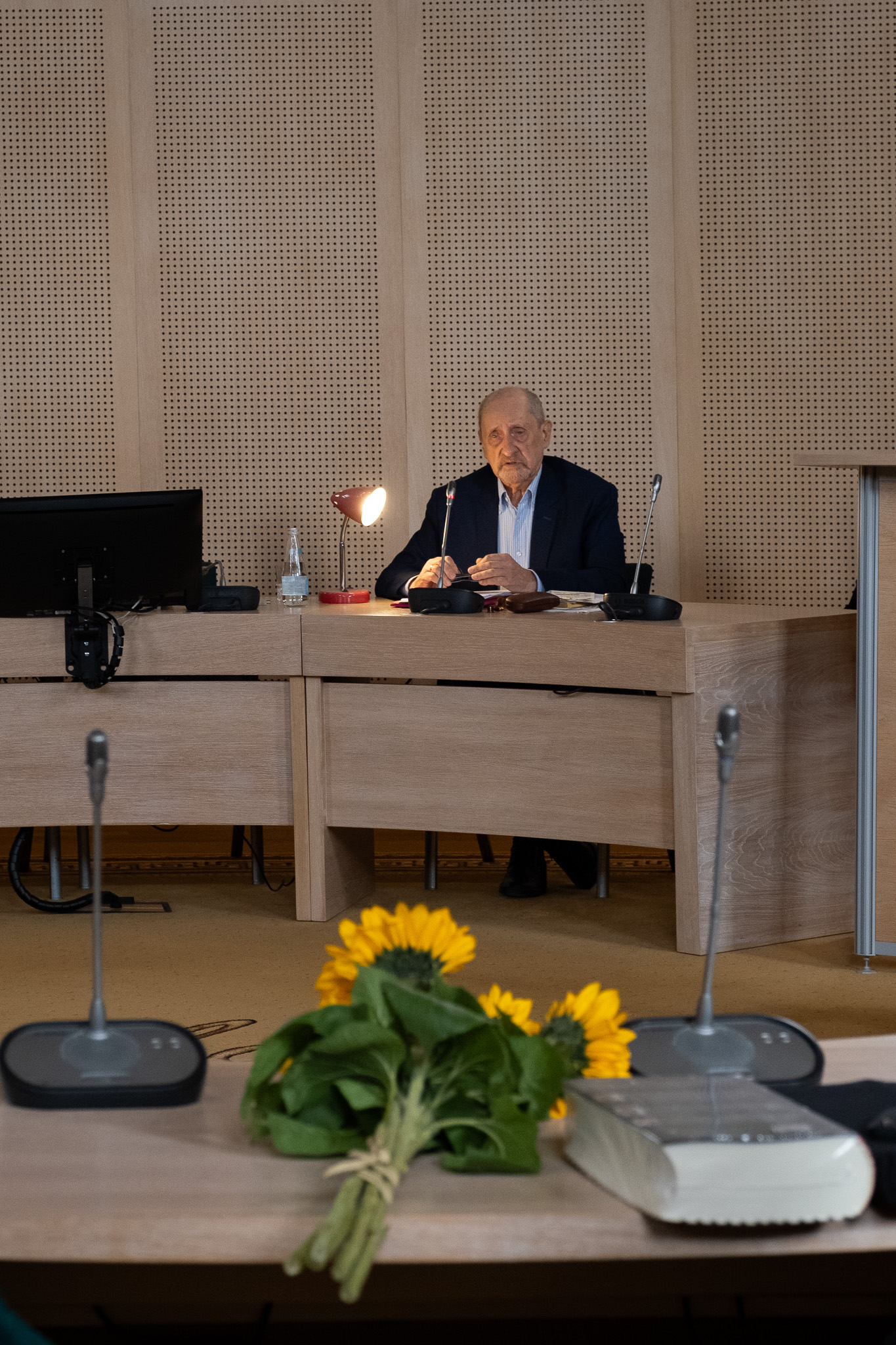 W Salonie Mickiewicza za stołem prezydialnym siedzi prof. Edward Balcerzan, obok niego zapalona lampa biurowa, przemawia do mikrofonu. Na pierwszym planie zdjęcia widać stół, na którym leży bukiet słoneczników.
