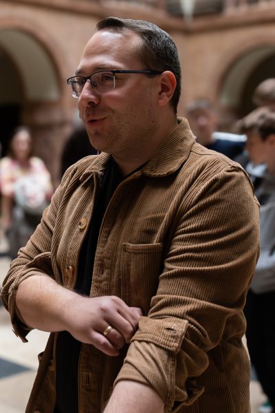 Przybliżone zdjęcie młodego mężczyzny w brązowej sztruksowej koszuli, czarnym t-shircie i okularach, skierowanego lekko profilem do obiektywu, mówiącego coś do rozmówców poza kadrem. Za nim rozmazane tło, w którym widać sylwetki ludzkie, ale nie widać ich twarzy.