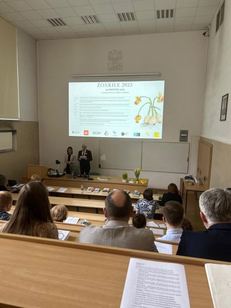 Zdjęcie z górnych rzędów ławek w sali Kuraszkiewicza, w których siedzą uczestnicy czytania, na dole wyświetla się prezentacja z programem wydarzenia, a przy mównicy stoi Dziekan WFPiK prof. Krzysztof Skibski i prodziekan ds. kształcenia i współpracy z otoczeniem dr Sylwia Karolak.