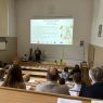 Zdjęcie z górnych rzędów ławek w sali Kuraszkiewicza, w których siedzą uczestnicy czytania, na dole wyświetla się prezentacja z programem wydarzenia, a przy mównicy stoi Dziekan WFPiK prof. Krzysztof Skibski i prodziekan ds. kształcenia i współpracy z otoczeniem dr Sylwia Karolak.