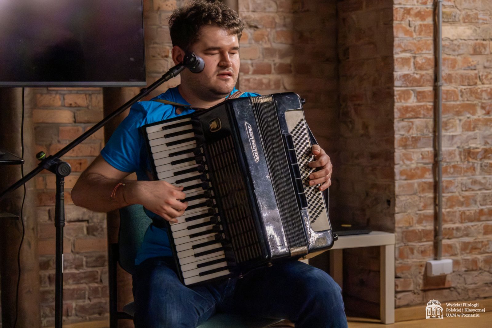 Mężczyzna z akordeonem siedzi w Sali Teatralnej i gra na akordeonie. Ma zamknięte oczy, jest niewidomy. Na jego twarzy widać skupienie.