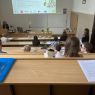 Zdjęcie z górnych rzędów ławek w sali Kuraszkiewicza, w których siedzą uczestnicy czytania, na dole wyświetla się prezentacja z programem wydarzenia, a przy mównicy stoi prof. Katarzyna Kuczyńska-Koschany,