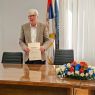 Starszy mężczyzna w jasnej marynarce stoi za stołem przybranym kwiatami, w ręku trzyma dyplom, za nim widać flagę Serbii i flagę Wydziału Filologicznego Uniwersytetu w Belgradzie.
