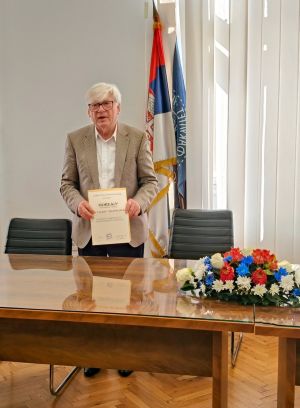 Starszy mężczyzna w jasnej marynarce stoi za stołem przybranym kwiatami, w ręku trzyma dyplom, za nim widać flagę Serbii i flagę Wydziału Filologicznego Uniwersytetu w Belgradzie.