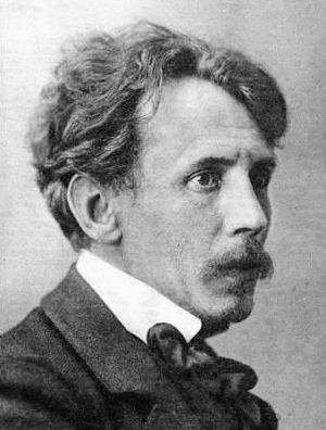 Czarno-biały portret Mikolajusa Konstantinasa Čiurlionisa w średnim wieku, przedstawiający mężczyznę z sumiastymi wąsami w wełnianym garniturze, białej koszuli i z czarną kokardą pod szyją.