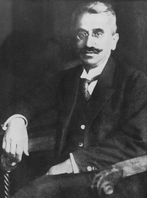 Czarno-białe zdjęcie prof. Stanisława Dobrzyckiego siedzącego na fotelu z ozdobnymi podłokietnikami. Profesor ma sumiaste wąsy, okrągłe binokle, jest ubrany w garnitur dawnego typu i koszule ze sztywnym, sterczącymi kołnierzykiem.