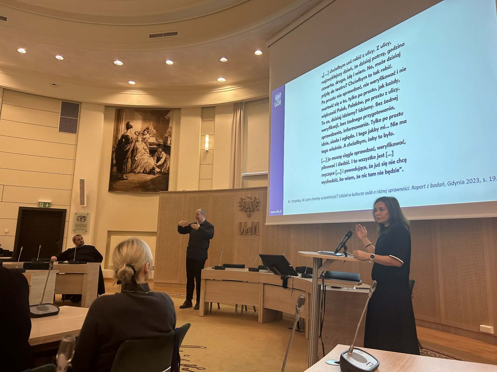 Prof. Karolina Ruta przemawia przy mównicy, prezentując coś na ekranie, towarzyszy jej tłumacz języka migowego.