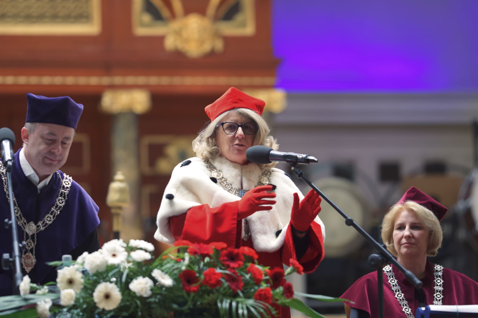 Rektor Bogumiła Kaniewska przemawia do mikrofonu, ubrana w czerwony płaszcz z gronostajami. Obok niej siedzi Prorektor Joanna Wójcik, a po drugiej stronie stoi Dziekan Tomasz Mizerkiewicz.
