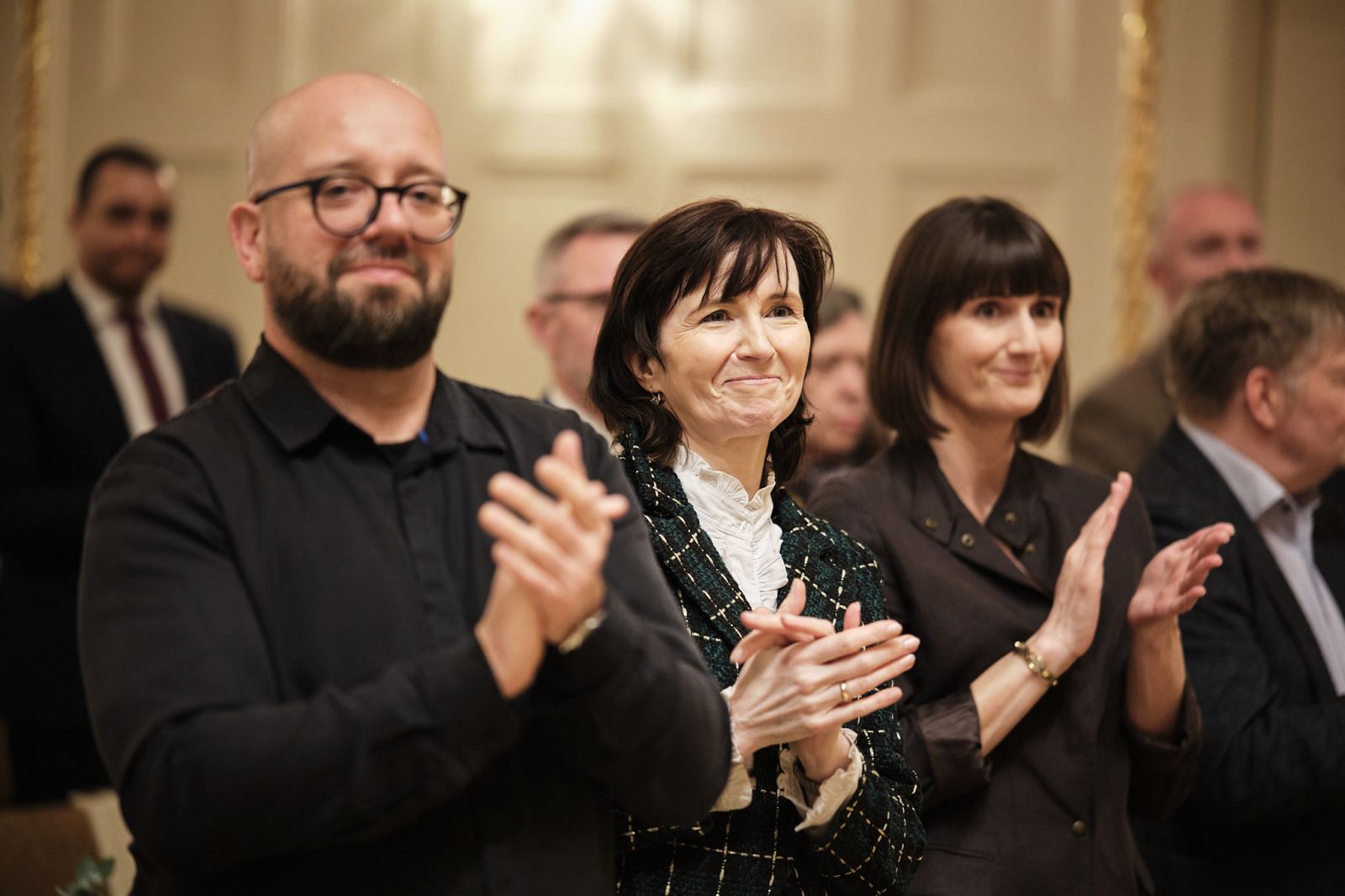 Prof. Marek Osiewicz, prof. Elżbieta Winiecka i mgr Natalia Czerwińska stoją w auli uniwersyteckiej i klaszczą, mają szerokie uśmiechy na twarzach.