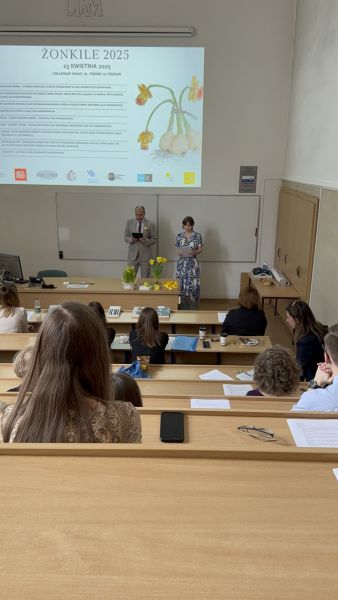 Zdjęcie z górnych rzędów ławek w sali Kuraszkiewicza, w których siedzą uczestnicy czytania, na dole wyświetla się prezentacja z programem wydarzenia, a przy mównicy stoi Prodziekan ds. współpracy międzynarodowej prof. Konrad Dominas i młoda kobieta, która trzyma mikrofon i czyta tekst z kartki.