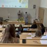 Zdjęcie z górnych rzędów ławek w sali Kuraszkiewicza, w których siedzą uczestnicy czytania, na dole wyświetla się prezentacja z programem wydarzenia, a przy mównicy stoi Prodziekan ds. współpracy międzynarodowej prof. Konrad Dominas i młoda kobieta, która trzyma mikrofon i czyta tekst z kartki.