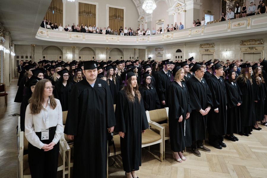 Uczestnicy absolutorium ubrani w togi i czapki absolwenckie siedzą w rzędach w Auli Uniwersyteckiej.