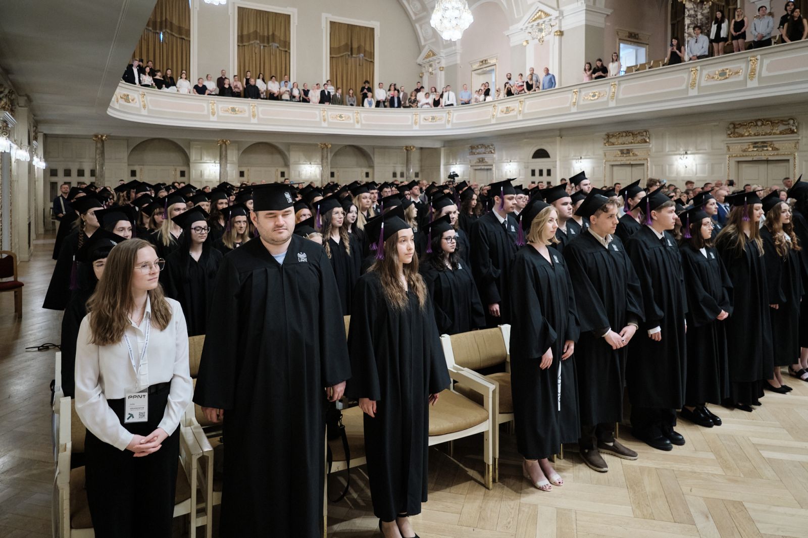 Uczestnicy absolutorium ubrani w togi i czapki absolwenckie siedzą w rzędach w Auli Uniwersyteckiej.