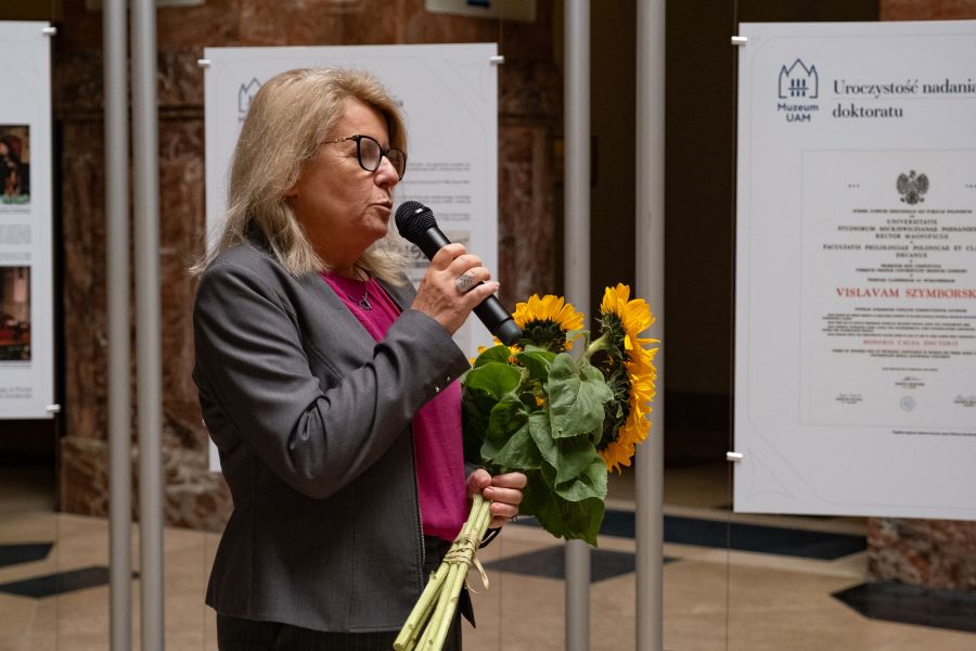 W holu głównym Collegium Maius, na tle tablic z wystawą o Wisławie Szymborskiej, stoi Jej Magnificencja Rektor UAM, przemawia do mikrofonu. W ręku trzyma bukiet słoneczników.