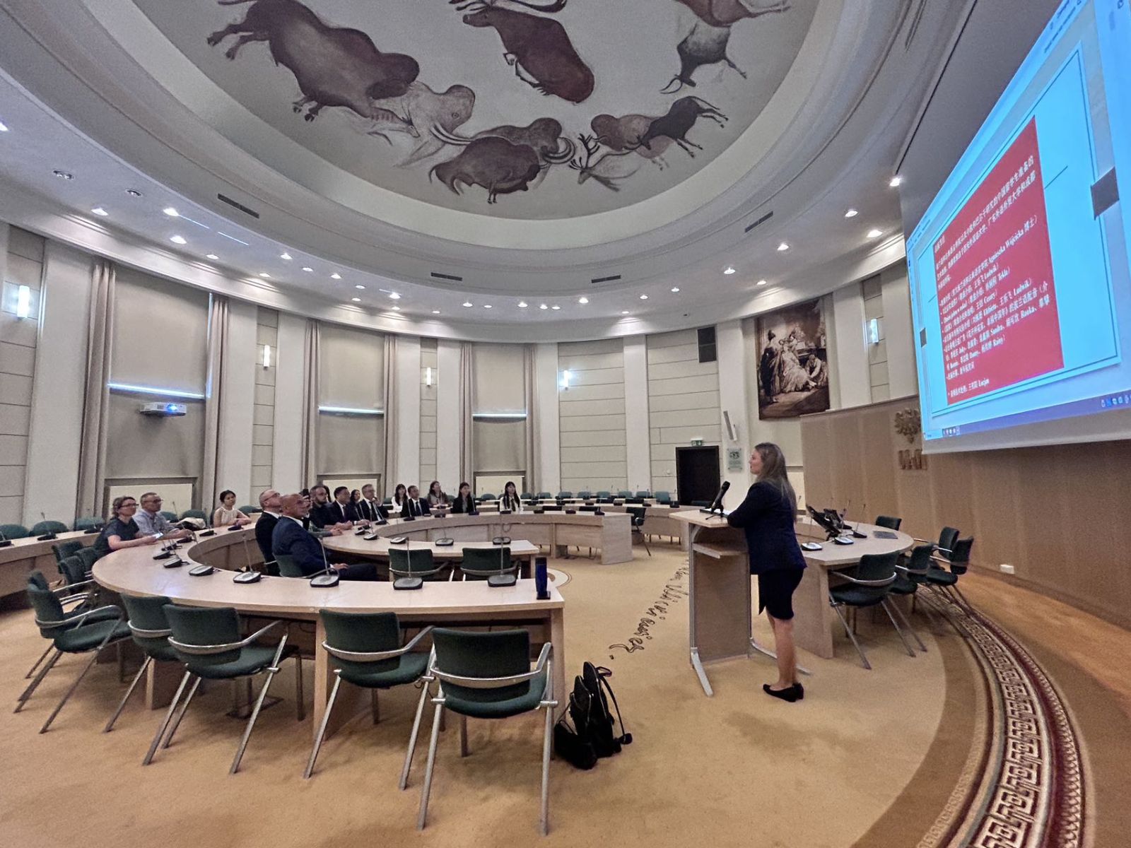 Widok Salonu Mickiewicza, przy mównicy stoi młoda kobieta, wygłaszając prezentację, na dużym ekranie wyświetla się jeden ze slajdów.