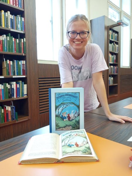 Młoda kobieta w okularach opiera się rękami o blat stołu, na którym stoją i leżą książki z kolorowymi ilustracjami.