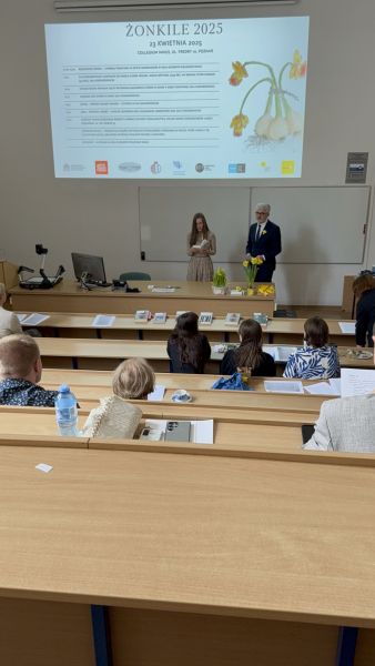 Zdjęcie z górnych rzędów ławek w sali Kuraszkiewicza, w których siedzą uczestnicy czytania, na dole wyświetla się prezentacja z programem wydarzenia, a przy mównicy stoi Prodziekan ds. nauki prof. Jerzy Borowczyk i młoda kobieta, która trzyma mikrofon i czyta tekst z kartki.