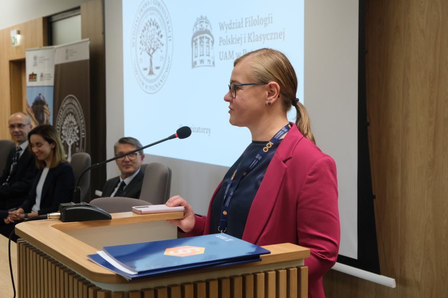 Kobieta w różowej marynarce i blond włosach związanych w kucyk przemawia do mikrofonu.