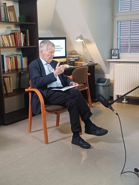 Prof. Jan Miodek siedzi w fotelu i przemawia, przed nim stoi mikrofon na statywie, a za nim regał z książkami oraz biurko z komputerem.