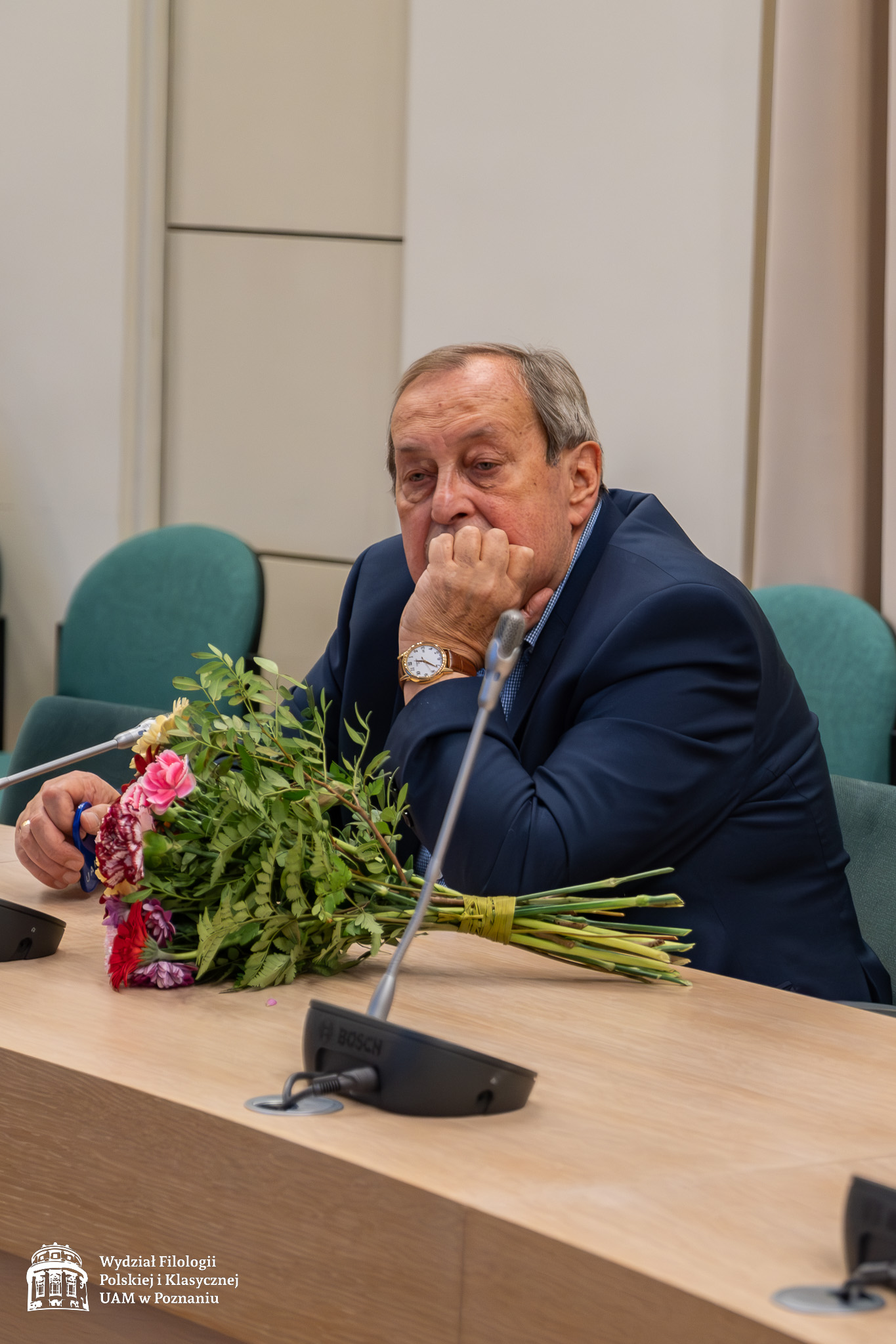 Starszy mężczyzna w garniturze siedzi wśród gości w Salonie Mickiewicza pod Kopułą, na stole przed nim leży bukiet kwiatów.