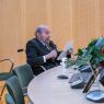 Prof. Tomasz Lewandowski siedzi w wózku inwalidzkim za stołem prezydialnym, jest ubrany w szary garnitur, w jednej ręce trzyma mikrofon, a w drugiej zadrukowaną kartkę, przemawia.