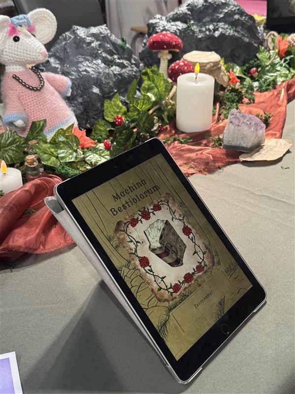 Na stole leży tablet z włączoną aplikacją do tworzenia fantastycznych chowańców, za tabletem widać kolorową instalację z roślinami, grzybami, skałami i świecą.