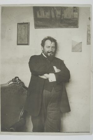 Sepiowy portret mężczyzny z brodą, ubranego w staroświecki surdut, opartego o ścianę, z założonymi na piersiach rękami.
